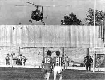 GAA Players take to the field amid British Army Activity in the base behind the goals.